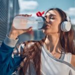 Eine Frau mit Over-Ear Bluetooth-Kopfhörern trinkt Wasser im freien