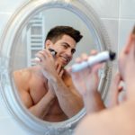 Mann rasiert sich mit elektrischen Rasierer vor einem Spiegel