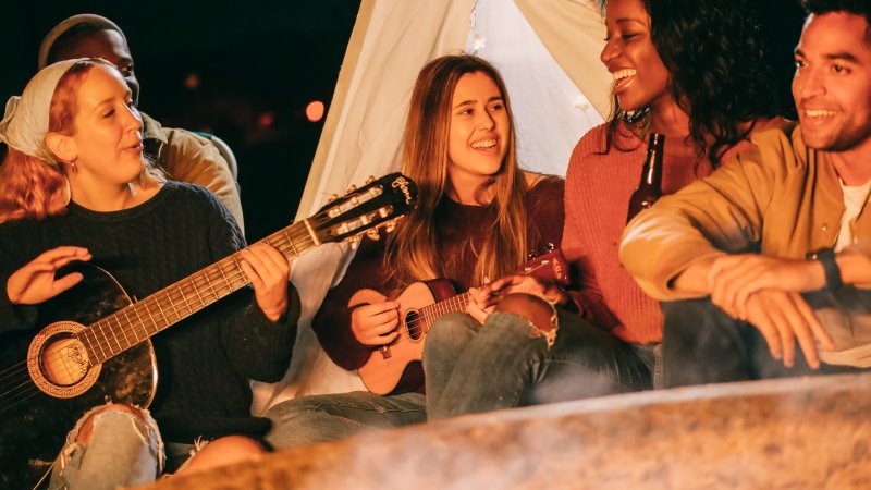 Eine Frau spielt am Lagerfeuer auf der Konzertgitarre Eine Gruppe sitzt am Lagerfeuer und singen während eine Freundin Konzertgitarre spielt