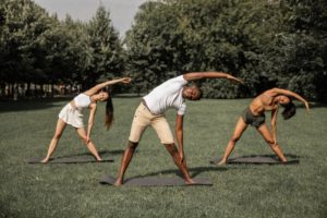 Zwei Frauen und ein Mann sind mit einem Sportshirt und einer Sporthose bekleidet. Es werden Dehnungsübungen auf einer Yogamatte in freier Natur ausgeführt.